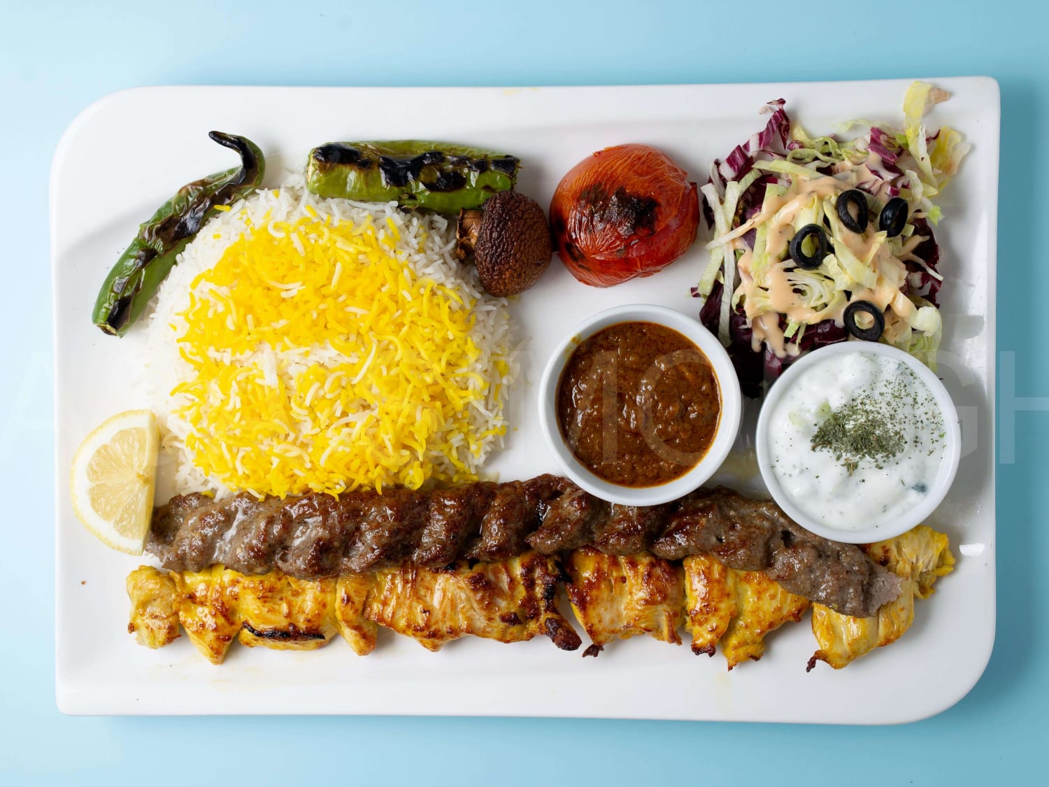 Eine Aufnahme von Soltani-Kebab auf blauem Hintergrund. Auf dem Teller sieht man Reis, ein Hähnchenspieß, ein Lammhackspieß, Salat und Tomatensoße.